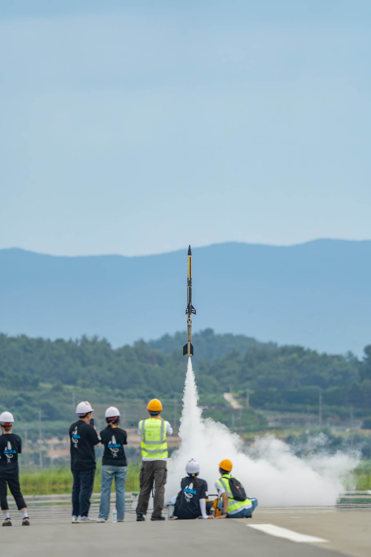 IDENTITY&sup2; climbing off the rail, smoke column vertical beneath it, team watching from the apron
